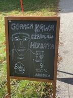 apron bartender cafe chalkboard closed_mouth clothes coffee cup ear grass hair hand happy holding_object internet_is_leaking irl logo millions_must_die photo poland polish_text sidewalk soyjak subvariant:chudjak_front traditional_media variant:chudjak wholesome // 3024x4032 // 1.7MB